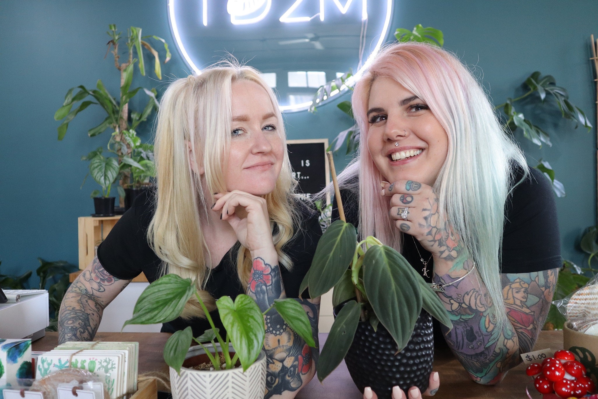 Talk Dirt 2 Me - Indoor Plant Store, Bec & Kathy the owners are very happy, looking at the camera showing their plants and pots. There is a big neon sign behind them